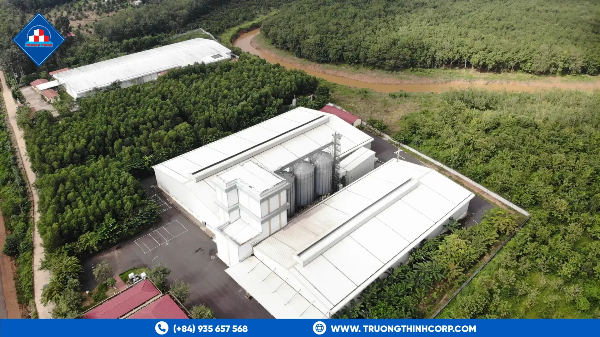 Aerial view of a feed processing plant with silos and steel-framed production halls surrounded by forest