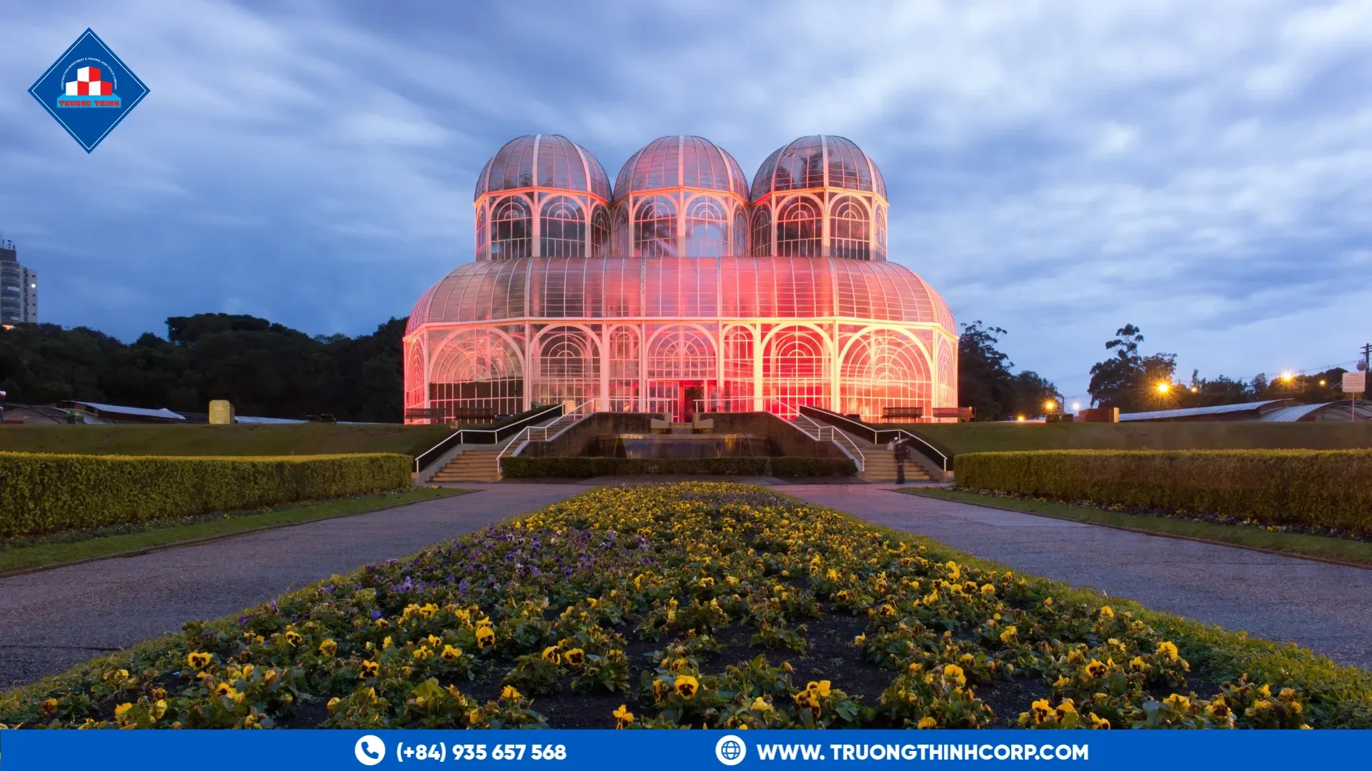 Large glasshouse with domes and a metal framework, lit at dusk above a garden
