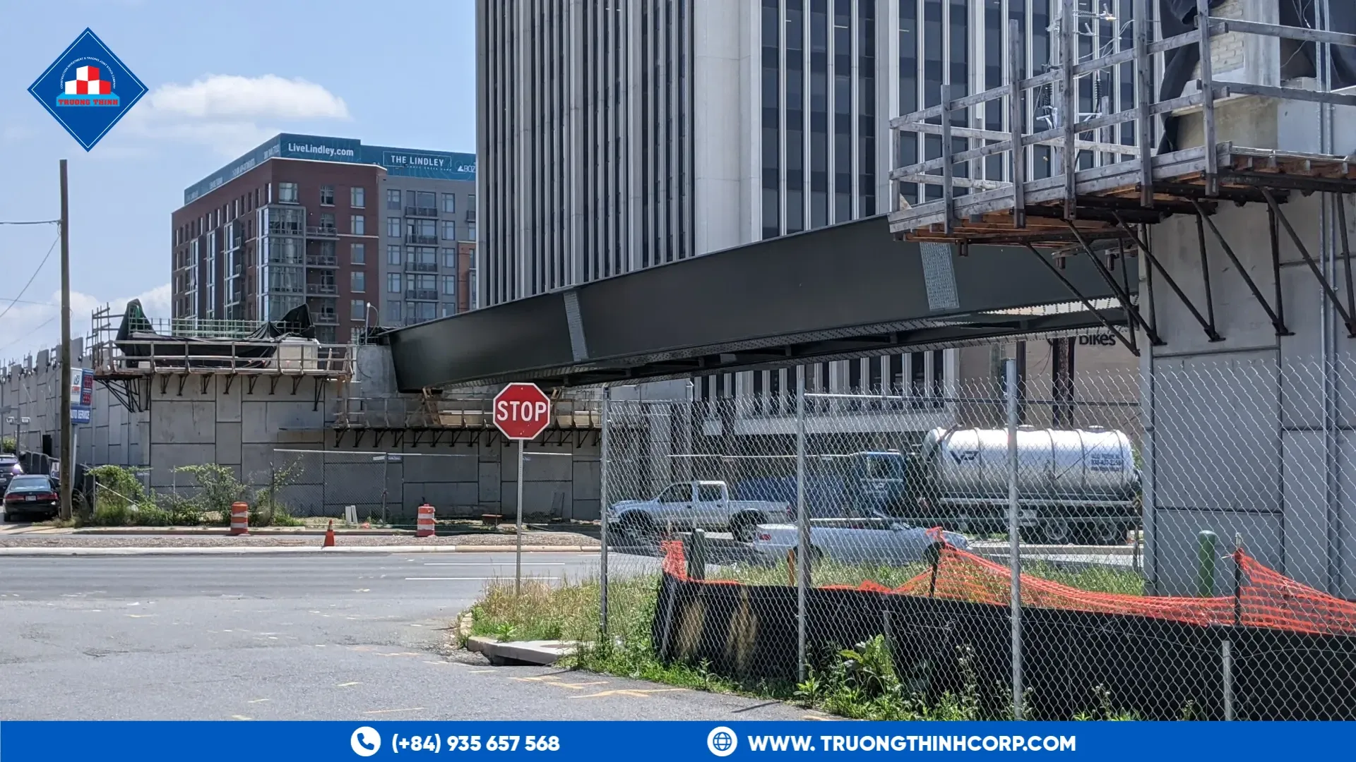 Steel girder spanning a road during bridge construction, with scaffolding and fencing beside a building